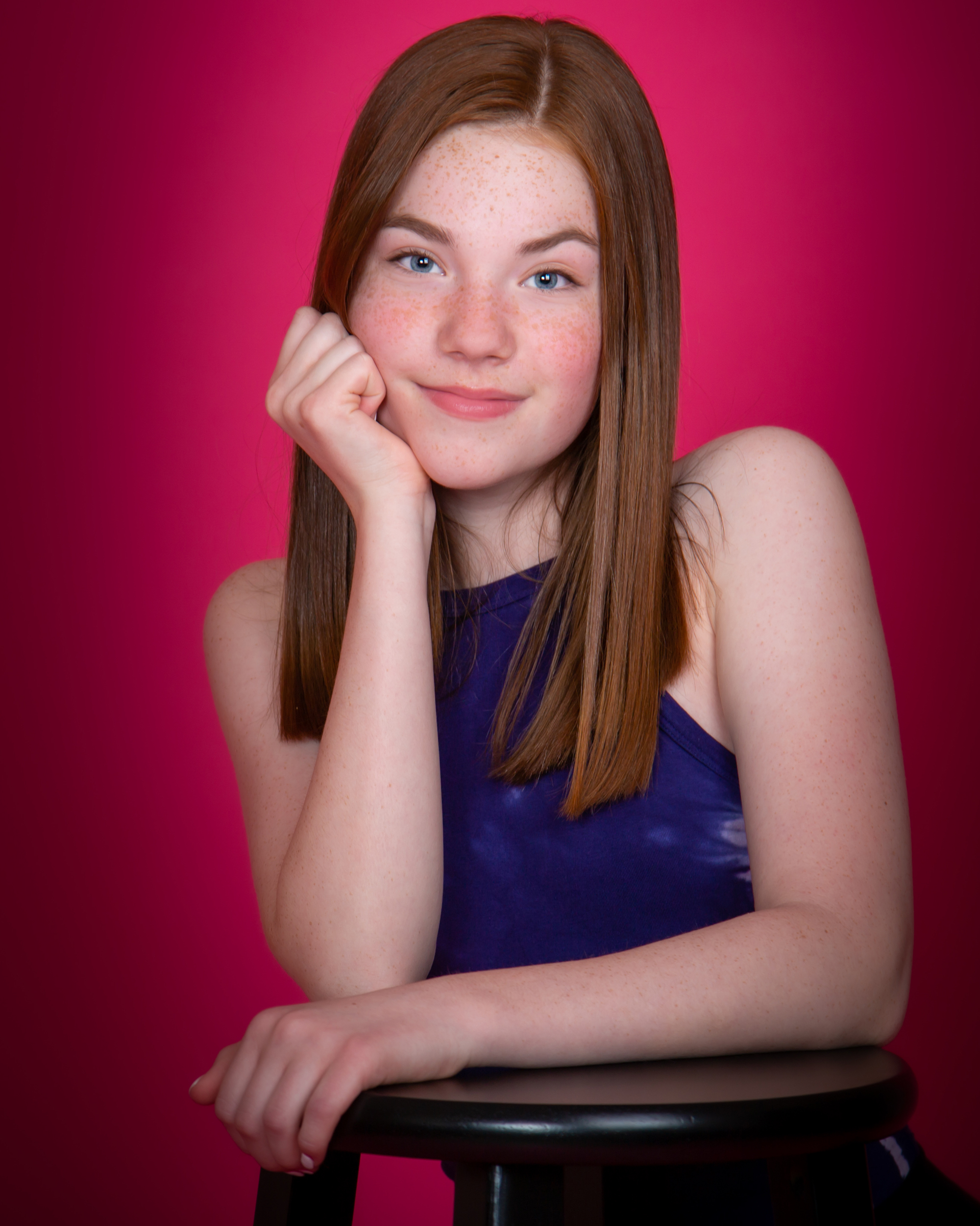 A young model is posed with her arms on a stool against a magenta background.